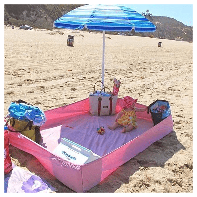 small child sitting in an improvised sand free play area on a sandy beach. The play area is made from a pink fitted bedsheet with a beach bag at each corner to hold up the sides. 