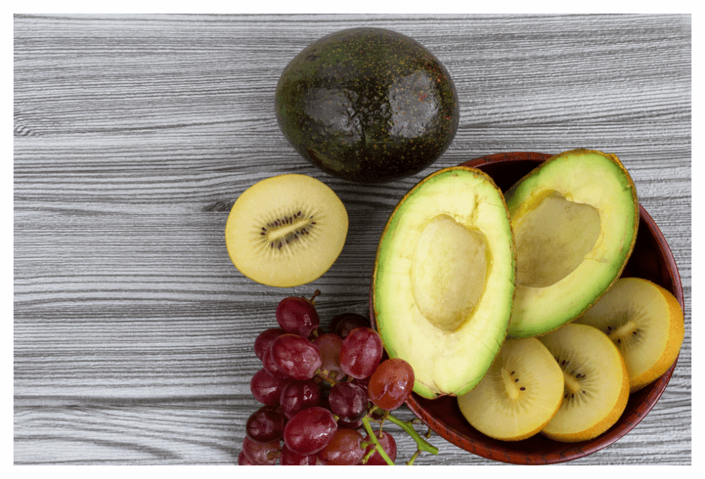 Still life of grapes, avocados and kiwi fruit against white and grey wooden table.