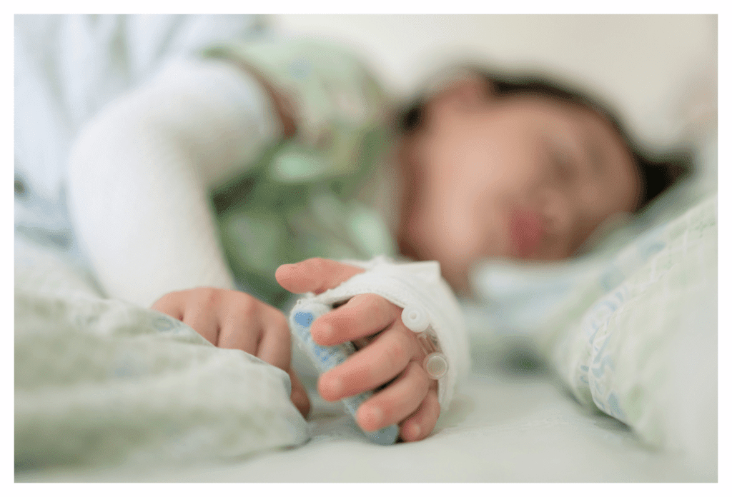 Asian girl sleeping in hospital bed. her arms are bandaged to protect her eczema and she has two ports in the back of her left hand.