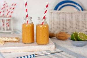 Eczema friendly papaya and banana smoothies, served in mini milk bottles with red and white striped paper straws. The orange smoothie contrasts with the blue and white background.