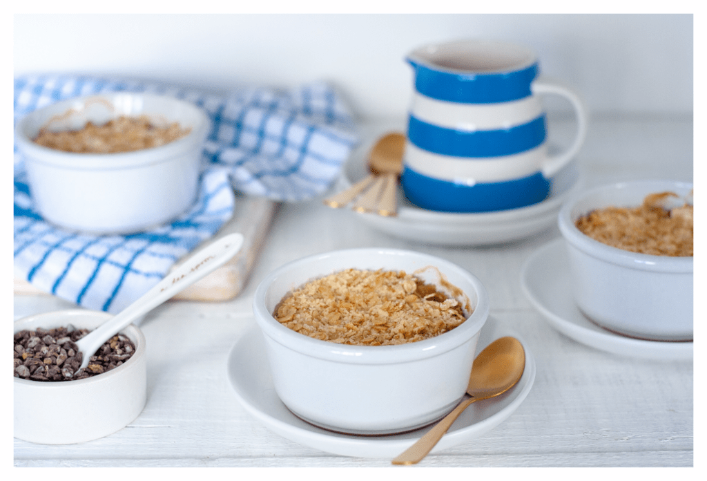 Eczema friendly pear oaty crumble desserts served in individual ceramic, white ramekins, with a side of carob nibs and classic blue and white striped jug of dairy free cream.