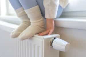 Young child sitting on a windowsill with her feet (in wooly socks) and hands resting on the radiator below. She has just turned up the thermostat.